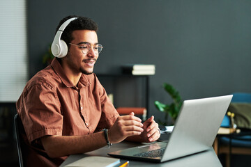 Young confident student in headphones communicating with online audience or teacher while sitting by desk in front of laptop - obrazy, fototapety, plakaty