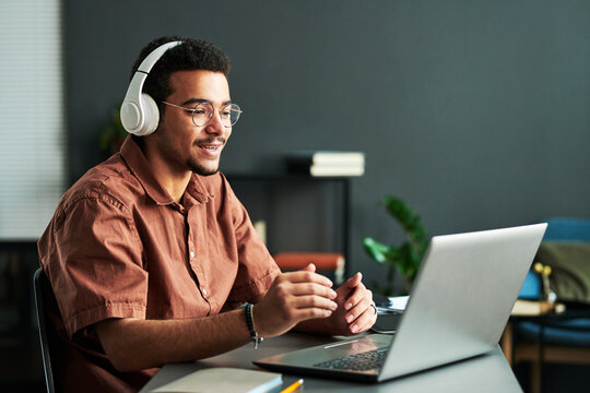 Young confident student in headphones communicating with online audience or teacher while sitting by desk in front of laptop
