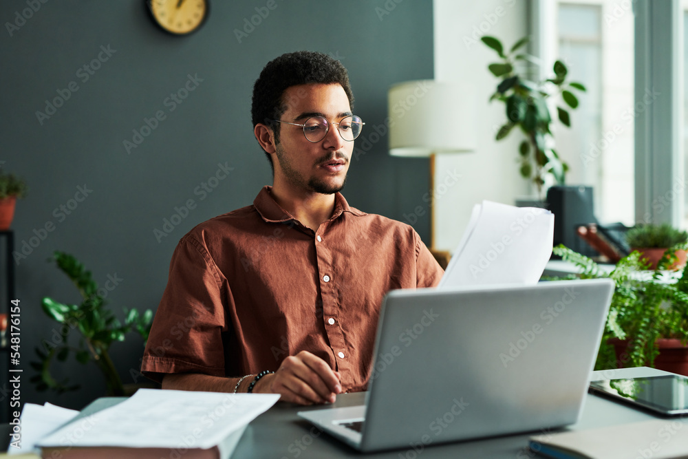 Poster young confident man with papers sitting by workplace in front of laptop and making report or explain