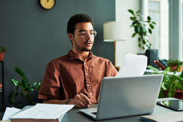 Young confident man with papers sitting by workplace in front of laptop and making report or...