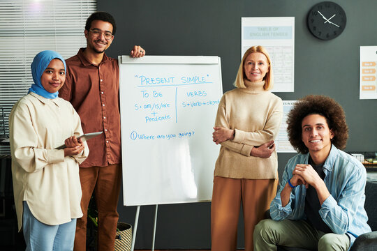 Several Happy Young Intercultural Students And Mature Blond Teacher Surrounding Whiteboard With Explanation Of Present Simple Tense