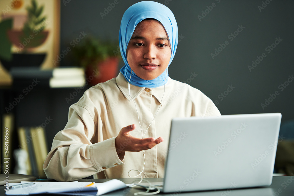 Poster young confident muslim woman in hijab taking part in webinar while sitting by desk in front of lapto