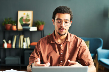 Young businessman or online student or tutor communicating with online audience while sitting by workplace in front of laptop - obrazy, fototapety, plakaty