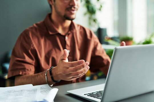 Focus on hand of young male tutor or student explaining something during online lesson while sitting by desk in front of laptop screen