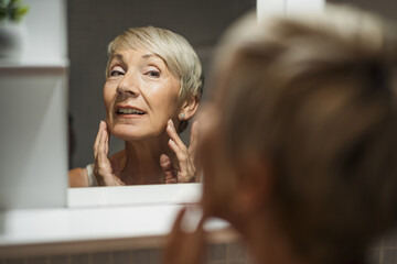 Mature woman looking at her face in the bathroom. She is examining wrinkles on her skin.
