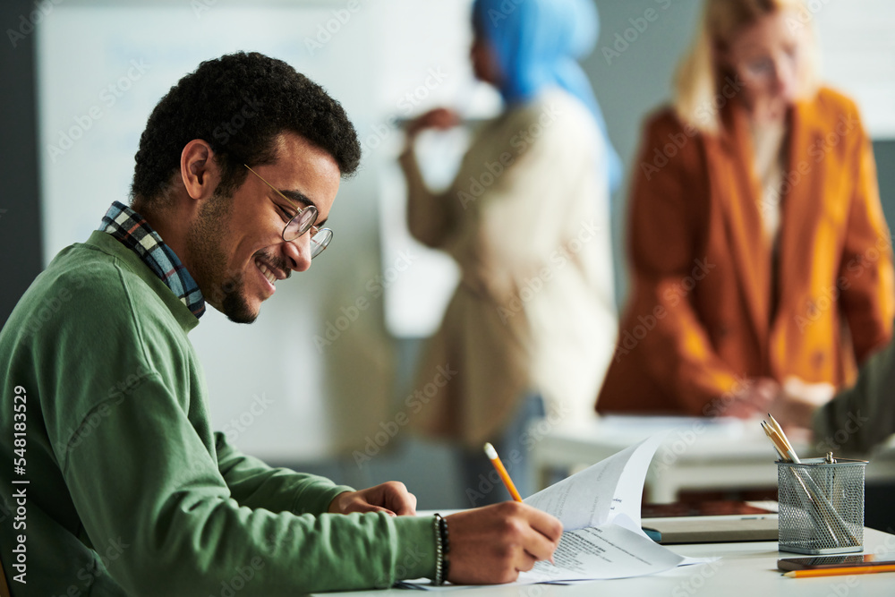 Poster Side view of happy Middle Eastern student putting ticks in front of right answers while carrying out grammar test by desk in classroom