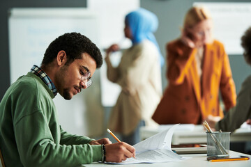 Young serious student in eyeglasses and casual pullover making notes on paper with exam text during individual work at lesson