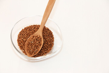 small grains of brown natural dry buckwheat in a transparent glass plate and in a wooden spoon