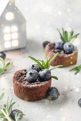 Chocolate brownies decorated with blueberries and rosemary with ceramic house on white textured background, bokeh lights. Christmas, New Year recipe concept. Close up