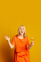 Party time. Thrilled young beautiful girl in orange dress tasting cocktail, having fun isolated over orange background. Vivid emotions, happiness, hobbies and joy