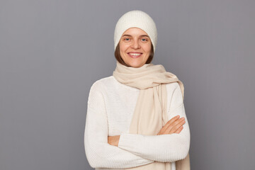 Horizontal shot of smiling delighted satisfied woman wearing white sweater, scarf and cap standing...