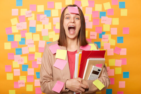 Image of funny crazy young adult woman holding papers, standing plastered with stickers and screaming loudly, posing against sticky notes for leaving reminder.