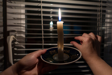 Blackout. A burning candle in the hands against the background of a window with blinds (close-up)