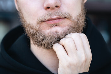 Close-up, a young man touches his beard with his hand.