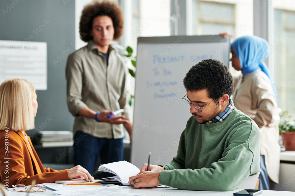 Poster young man making notes in copybook while sitting by desk at lesson of english grammar against guy ex