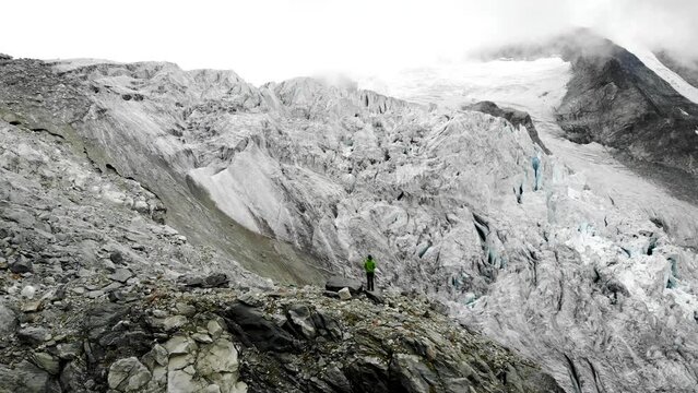 Aerial flyover over a hiker towards the edge of the Moiry glacier near Grimentz in Valais, Switzerland with a view of the icy crevasses and alpine peaks hidden in clouds