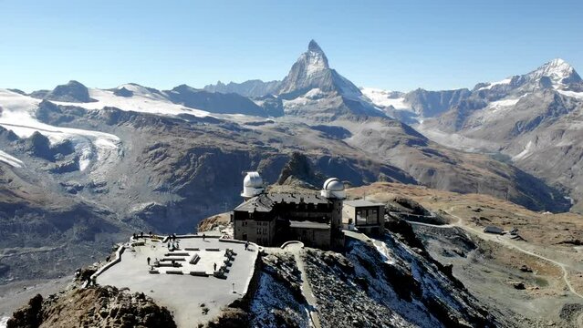 Aerial flyover over Gornergrat in Zermatt, Switzerland with a view of the Matterhorn and a train departing the train station revealed behind the observatory.