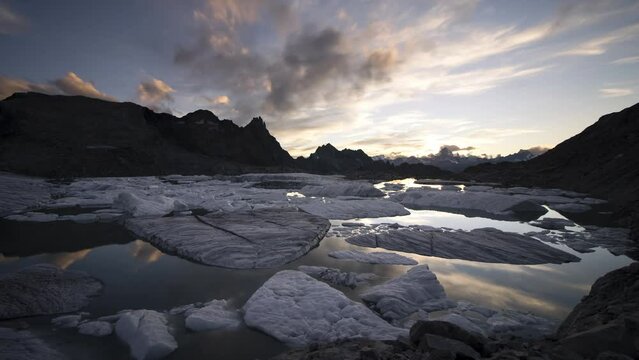 Day to night sunset time-lapse of a lake full of icebergs from a melting glacier in the remote Swiss Alps as the sky changes from glowing to full of stars before being engulfed by clouds and darkness