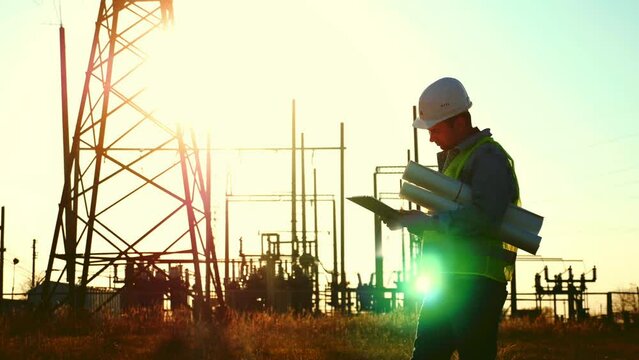 Silhouette of engineer walk on field with electricity towers. Electrical engineer with high voltage electricity pylon at sunset background. Power workers at work concept.