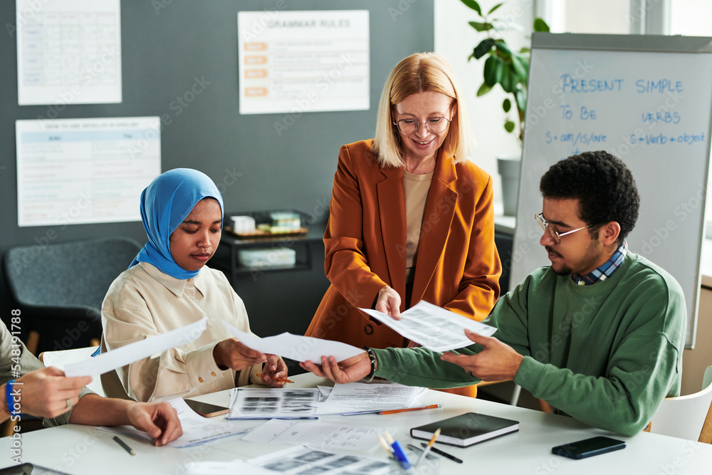 Poster Group of intercultural students taking grammar tests while teacher passing papers to them at lesson of English language