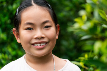 Portrait of Asian child girl in 8 to 9 years old, close up to face, smiling, looking at camera, outdoor image, blurred background of green plant garden.