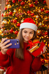 Cheerful young girl in santa hat, making selfie with gift box near Christmas tree. Winter holidays happytime, cozy warm atmosphere, sharing presents and good moments online