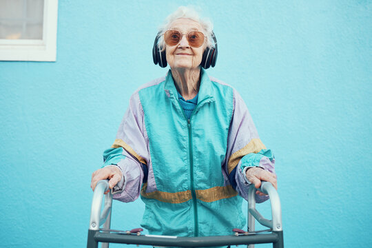City, fashion and senior woman with headphones listening to music, audio and radio on blue background wall. Freedom, style and elderly female with modern, urban and trendy clothes with walker in town
