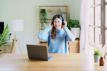 Portrait of a teenage Asian woman using a computer, wearing headphones to study online via video conferencing on a wooden desk at home