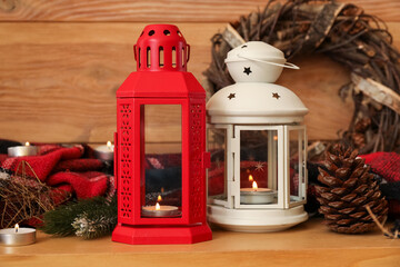 Christmas lanterns with burning candles, wreath and plaid on table near wooden wall