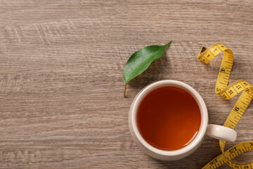 Cup of herbal diet tea and measuring tape on wooden table, flat lay with space for text. Weight loss concept