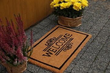Doormat with word Welcome and beautiful plants on paving stone near entrance
