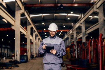A heavy industry worker is scrolling on tablet and finishing checklist while standing in facility.