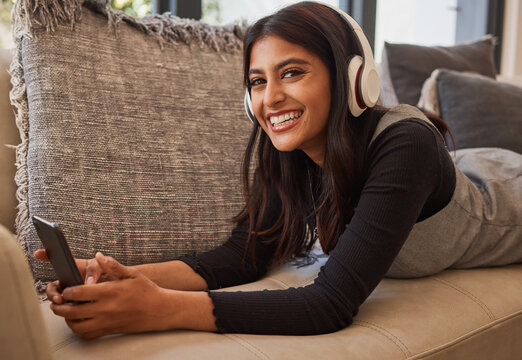 Woman, happy and headphone phone music of a person listening to a podcast or video watching at home. Portrait of an indian person with happiness and smile about mobile technology on living room sofa