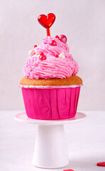 Cupcake with pink icing decorated with sugar sprinkles and heart topper on a small cake stand....
