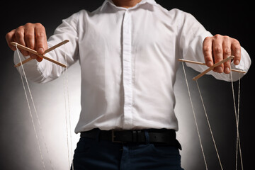 Man in formal outfit pulling strings of puppet on dark background, closeup