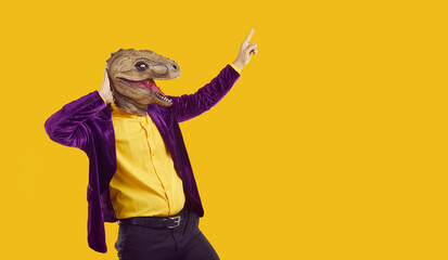 Eccentric funny showman in rubber dinosaur mask and velvet jacket pointing index finger on copy space on yellow background. Party, masquerade with costumes. Funky dancing guy using animal style. © Studio Romantic