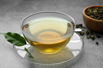 Fresh green tea in glass cup with saucer and leaves on grey table, closeup