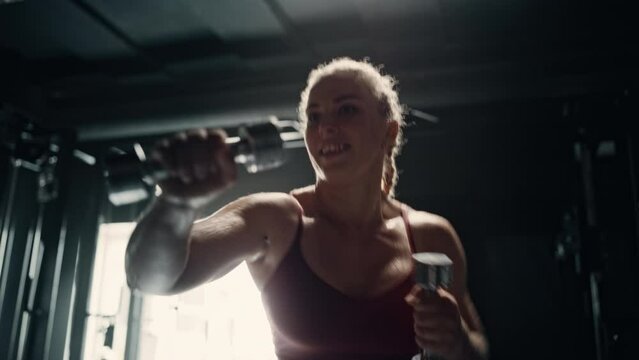 A Professional Female Weightlifter Doing her Early Morning Exercises in an Empty Hardcore Gym. A Fit Young Woman Smiling Happily While Working Out and Training Using Dumbbells. Arc Slow Motion Shot