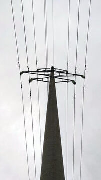 A tall concrete pole with electrical wires. A cement pylon of a power line under a light gray sky that is covered with clouds. Oprah is holding several high voltage metal wires.