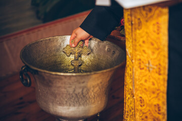 A priest is holding a golden cross necklace and purifying it in water for an orthodox baby child...