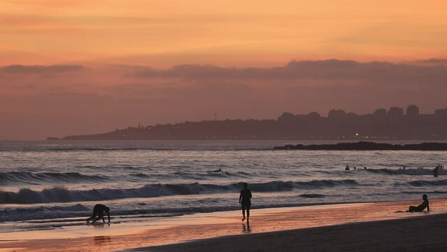 Silhouettes of people walking on sandy beach and surfers at the sunset background on the Atlantic coast of Portugal.