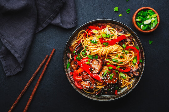 Vegan stir fry egg noodles with vegetables, paprika, mushrooms, chives and sesame seeds in bowl. Asian cuisine dish. Black table background, top view