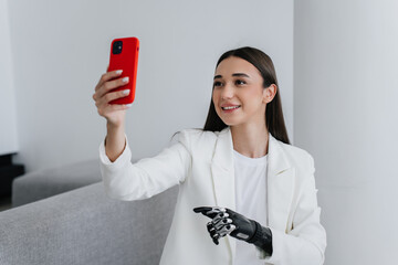 European brunette girl with bionic artificial hand in white suit holds phone, making video call by...