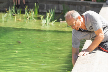 Senior man in glasses sitting by fountain in the city park of Prague, Czech Republic. Throws a coin into the pond water.