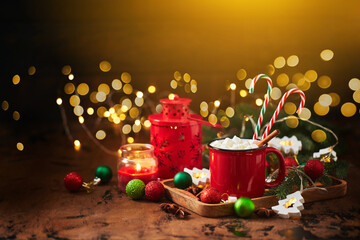 Red cup of hot cocoa with marshmallows and candy canes on wooden table with christmas tree and glowing garland for christmas