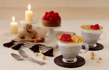 Chia pudding dessert. White wooden table with 2 glass cups of chia pudding with crushed hazelnuts and fresh raspberries and candle lights. High angle of view, no people.