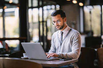 Focused young businessman is typing on a laptop while working remotely from coffee shop.