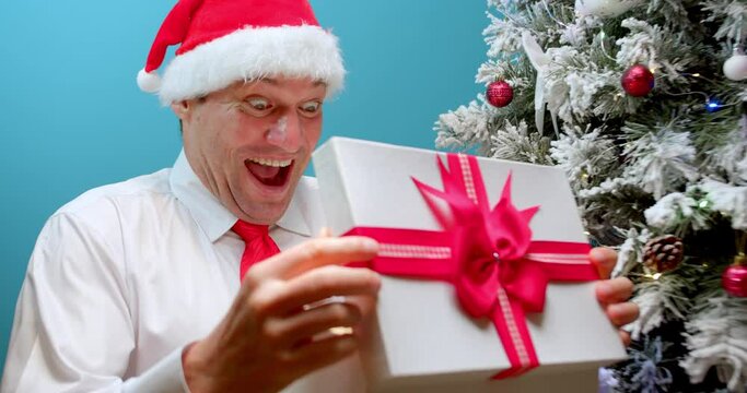 Happy smiling dad in Christmas hat Santa Claus receives a gift, rubs his hands before opening box with a gift, looks into box with surprise, happy face man delighted with surprise opens box with gifts