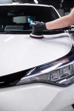 Employee of a car wash or a car detailing studio polishes the paintwork of a white car with an electric polisher