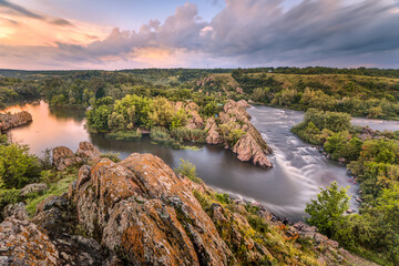 Beautiful summer sunset at the river with orange sky, clouds, rocks, green trees, flowers and water flow with reflection. Ukraine nature water background - 548228563